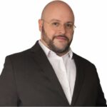 Portrait de Julien Riposo, photographié dans un cadre professionnel. Il apparaît en tenue élégante, le regard assuré, avec une expression calme et concentrée. La lumière met en valeur les traits de son visage et souligne une posture droite et confiante. L’arrière‑plan, sobre et légèrement flouté, renforce l’attention portée au sujet et donne à l’image une tonalité moderne et institutionnelle.