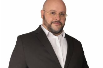 Portrait de Julien Riposo, photographié dans un cadre professionnel. Il apparaît en tenue élégante, le regard assuré, avec une expression calme et concentrée. La lumière met en valeur les traits de son visage et souligne une posture droite et confiante. L’arrière‑plan, sobre et légèrement flouté, renforce l’attention portée au sujet et donne à l’image une tonalité moderne et institutionnelle.