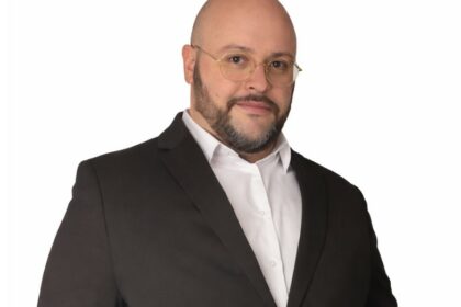 Portrait de Julien Riposo, photographié dans un cadre professionnel. Il apparaît en tenue élégante, le regard assuré, avec une expression calme et concentrée. La lumière met en valeur les traits de son visage et souligne une posture droite et confiante. L’arrière‑plan, sobre et légèrement flouté, renforce l’attention portée au sujet et donne à l’image une tonalité moderne et institutionnelle.