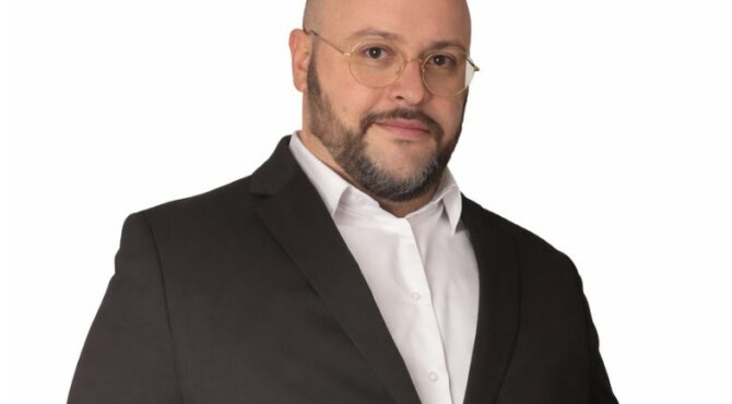 Portrait de Julien Riposo, photographié dans un cadre professionnel. Il apparaît en tenue élégante, le regard assuré, avec une expression calme et concentrée. La lumière met en valeur les traits de son visage et souligne une posture droite et confiante. L’arrière‑plan, sobre et légèrement flouté, renforce l’attention portée au sujet et donne à l’image une tonalité moderne et institutionnelle.