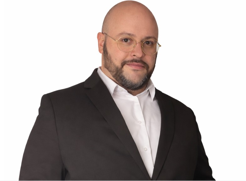 Portrait de Julien Riposo, photographié dans un cadre professionnel. Il apparaît en tenue élégante, le regard assuré, avec une expression calme et concentrée. La lumière met en valeur les traits de son visage et souligne une posture droite et confiante. L’arrière‑plan, sobre et légèrement flouté, renforce l’attention portée au sujet et donne à l’image une tonalité moderne et institutionnelle.