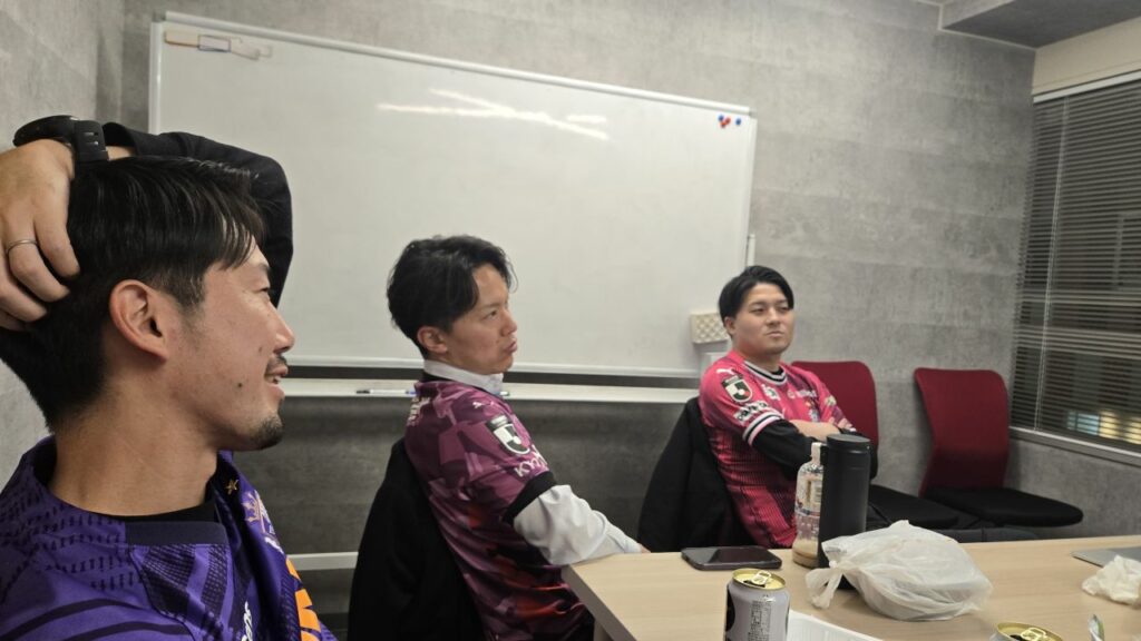 Photo de supporters réunis autour d’un écran diffusant un match de J‑League, partageant repas et analyses dans une ambiance conviviale.