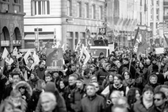 Une silhouette de l’hémicycle vue en plongée, traversée par une fissure lumineuse qui sépare symboliquement les blocs politiques. L’atmosphère est crépusculaire, évoquant la fin d’un cycle républicain et la recomposition des frontières du légitime.
