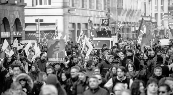 Une silhouette de l’hémicycle vue en plongée, traversée par une fissure lumineuse qui sépare symboliquement les blocs politiques. L’atmosphère est crépusculaire, évoquant la fin d’un cycle républicain et la recomposition des frontières du légitime.