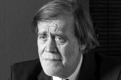 Jean‑Pierre Servel, assis dans son bureau à Carqueiranne, entouré de sa bibliothèque de livres anciens, regard bleu et expression concentrée.