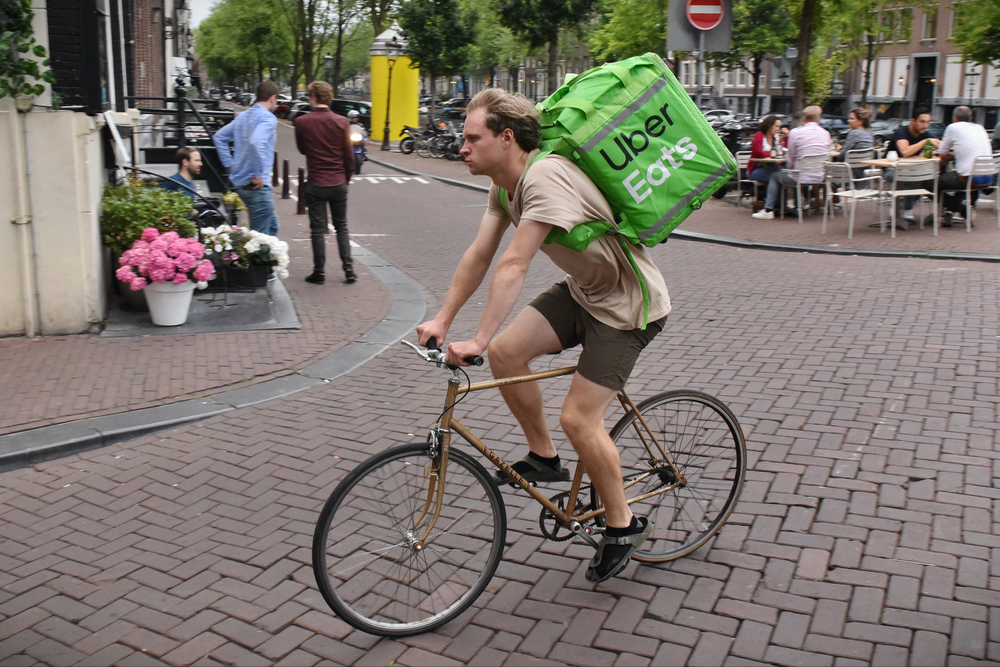 Livreurs Uber Eats et Deliveroo circulant en ville, illustrant la plainte déposée pour traite d’êtres humains et les conditions de travail dénoncées.