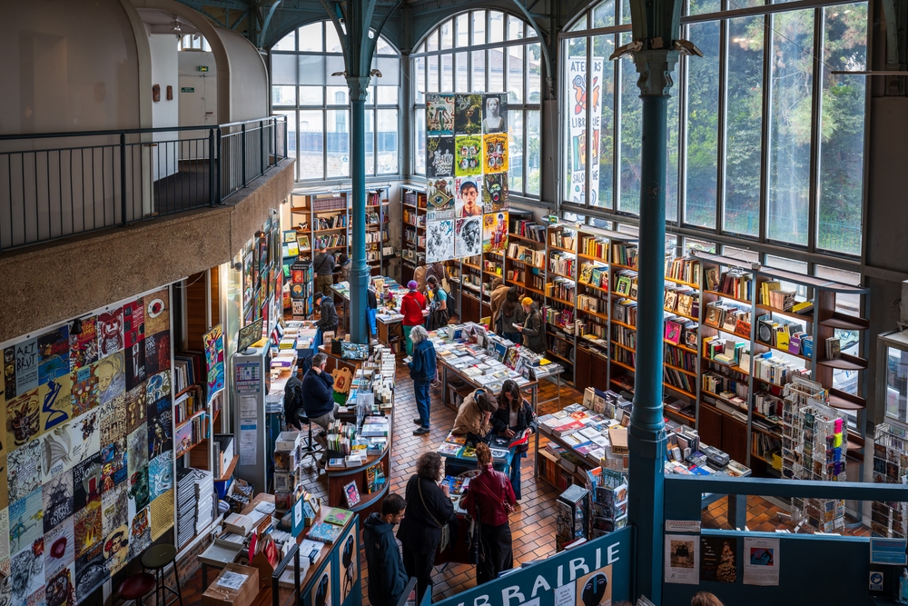 Une librairie indépendante animée, avec des lecteurs parcourant les rayons et un libraire en discussion avec ses clients.